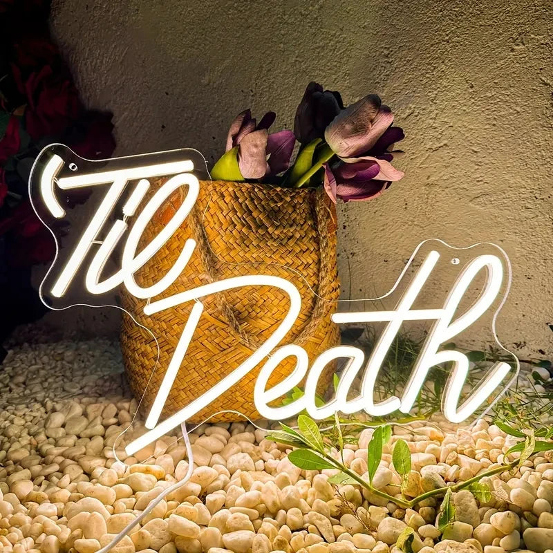 Neon sign with 'Til Death' text in front of a woven basket with flowers on a textured surface.