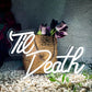 Neon sign with 'Til Death' and a woven basket with flowers on pebbles.